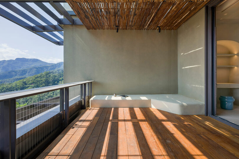 A modern balcony with wooden flooring, a built-in white bench, partial bamboo roof, and mountain views in the background.
