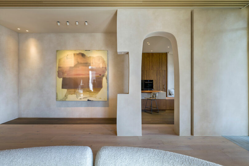 Modern minimalist living space with beige walls, abstract artwork, an arched doorway, and a view into a wooden-finished kitchen area with a bar stool.