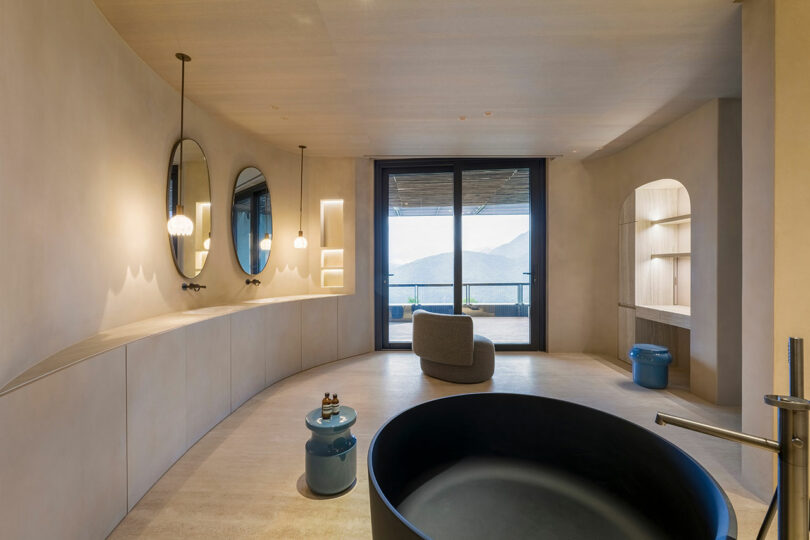 Modern minimalist bathroom with curved beige walls, two round mirrors above a long counter, freestanding black tub, blue side tables, and a large window overlooking mountains.