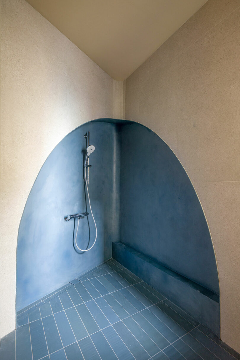 Modern shower with a curved blue alcove, handheld showerhead, and matte blue floor tiles. Walls and ceiling are light beige.