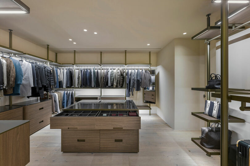 Modern walk-in closet with neatly organized clothing on racks, wooden drawers and shelves, and accessories displayed on open shelving and in glass-topped drawers.