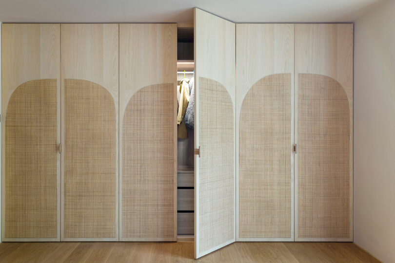 A built-in wooden wardrobe with light wood and woven rattan doors, one of which is open to reveal hanging clothes and interior drawers.