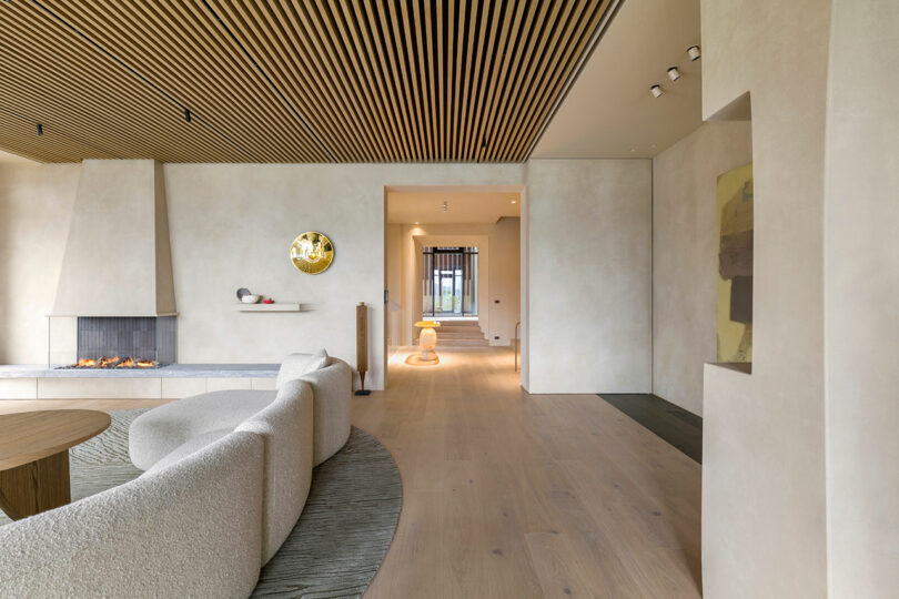 Modern living room with a curved sofa, round coffee table, gas fireplace, wooden slat ceiling, and minimalist decor. A hallway with a lamp is visible in the background.