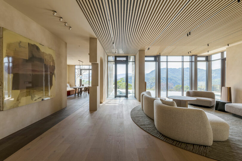 Modern living room with curved sofas, round coffee tables, wood flooring, large abstract art, and floor-to-ceiling windows overlooking a scenic mountain view.