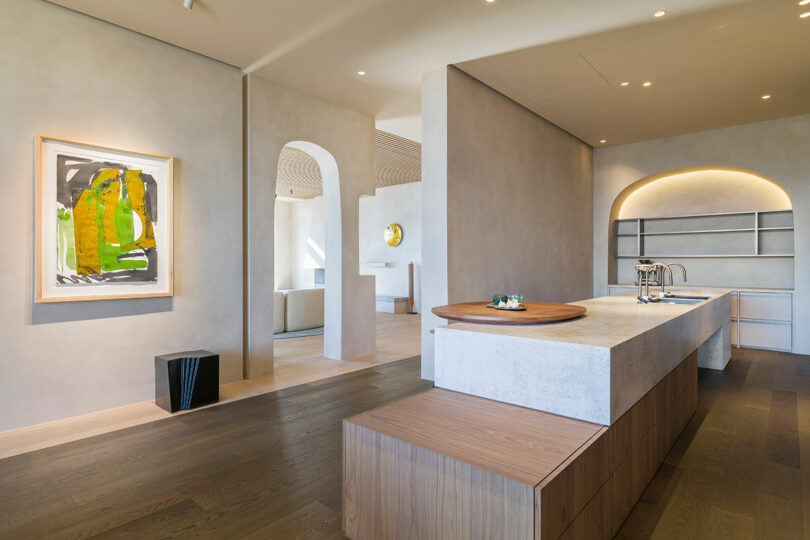 Modern kitchen with a minimalist island, light wood floors, neutral walls, built-in shelves, abstract artwork, and soft, recessed lighting.