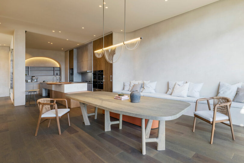 Modern open-plan kitchen and dining area with wooden table, chairs, built-in bench seating, minimalist lighting, and neutral-toned decor.