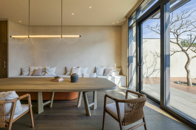 A modern dining area with a wooden table, chairs, neutral-toned sofa with cushions, large windows, and natural light streaming in.