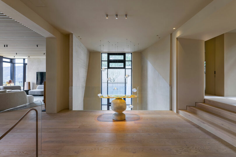 Modern interior hallway with light wood flooring, a sculptural centerpiece, large windows, and neutral-toned walls, leading to a living area and staircase.