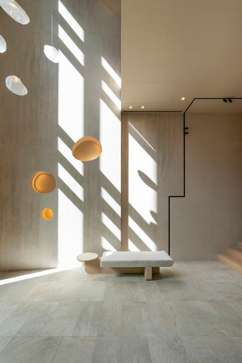 Minimalist interior with light gray walls, geometric shadows, built-in seating, circular wall accents, and hanging glass fixtures. Natural light streams in from an unseen window.