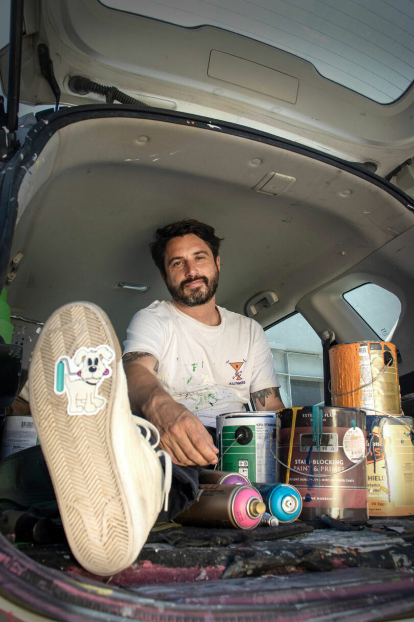 David Kaul sits in the open trunk of a car, surrounded by paint cans and spray bottles, one foot forward showcasing a shoe with a dog sticker on the sole.