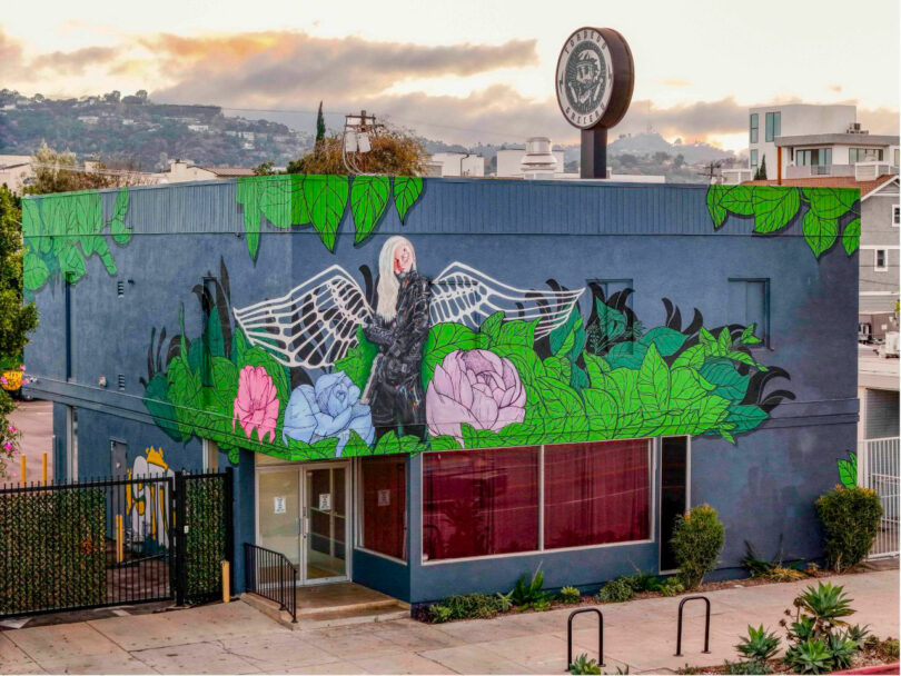 A blue building features a mural by David Kaul of a woman with white wings, surrounded by large green leaves and colorful flowers; a circular sign is visible on the roof.
