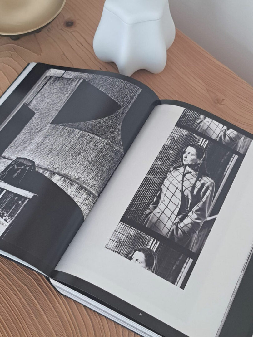 An open book on a wooden surface shows black and white photographs of a woman standing next to a wire fence and textured concrete wall. Nearby, a lamp and white ornament reflect the minimalist style of Dorothée Boissier.