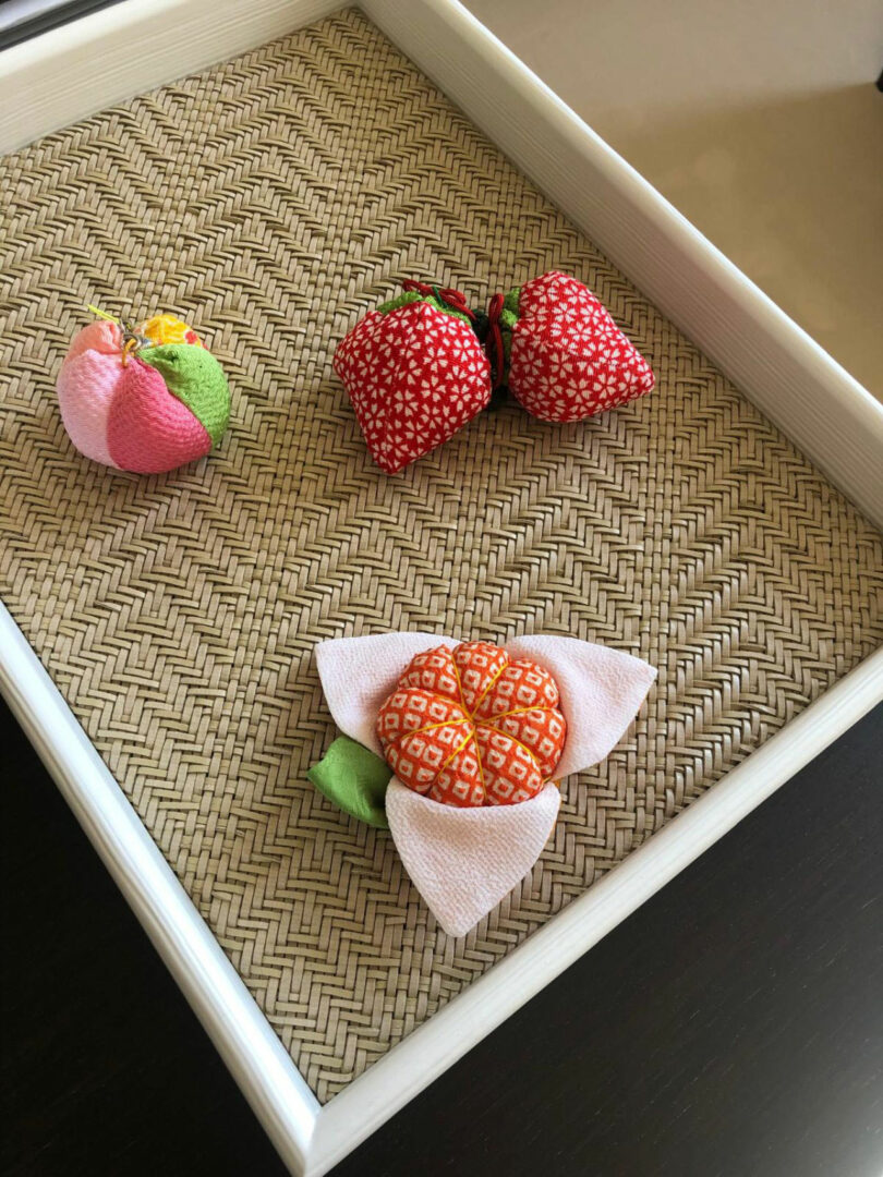 Four fabric fruits, including strawberries and a peach, are displayed on a woven tray inside a white rectangular frame, reflecting the refined aesthetic inspired by Dorothée Boissier.