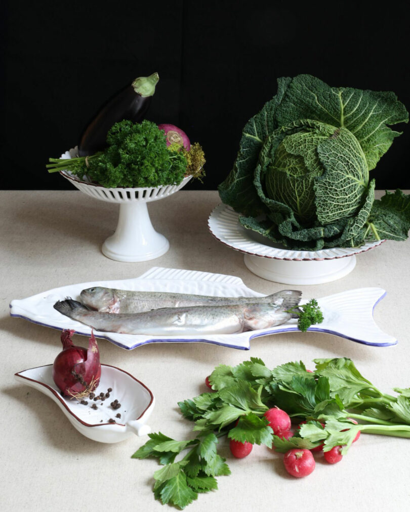 A whole raw fish on a fish-shaped plate is surrounded by cabbage, eggplant, parsley, red onion, celery, radishes, and a dish with peppercorns in a style reminiscent of Dorothée Boissier’s elegant culinary presentations.