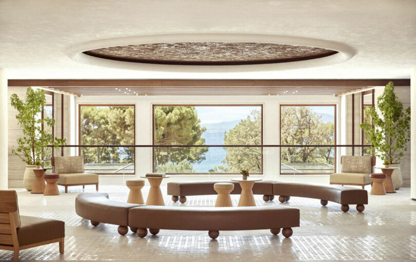 A modern lobby designed by Dorothée Boissier features curved beige sofas, small round tables, large windows, and lush greenery, offering tranquil views of trees and water outside.