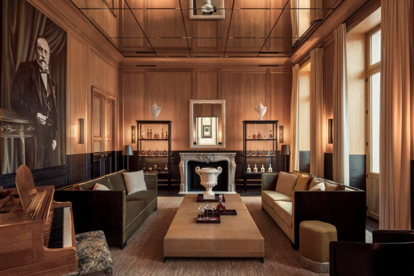 Elegant living room with wood paneling, a large portrait, and Dorothée Boissier-inspired design. Two sofas face a central table, a piano graces the space, and symmetrical cabinets flank a fireplace topped with a mirror. Natural light floods in from tall windows.