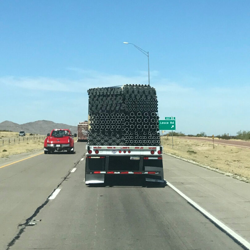 A truck carrying stacked black pipes drives on a highway; a red pickup follows behind. A green road sign for "Lasco Rd" is visible on the right.