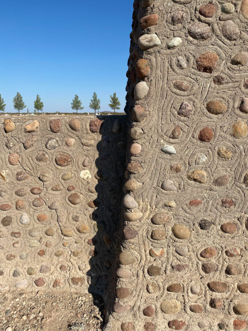 Close-up of a stone and concrete wall casting a shadow, with a row of evenly spaced trees visible in the background under a clear blue sky.