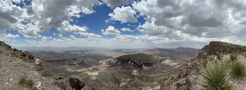 Wide panoramic view of a desert landscape with rocky cliffs, scattered vegetation, and partly cloudy sky; two people are visible on opposite sides of the image.