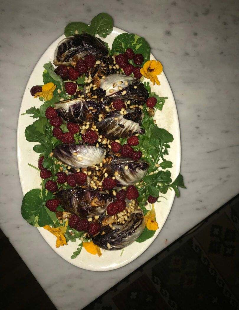 Oval platter with grilled radicchio, fresh raspberries, greens, pine nuts, and edible yellow flowers on a marble surface.