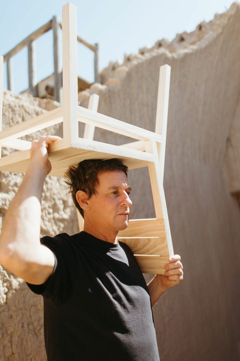 A man in a black shirt holds an upside-down wooden chair on his head while standing outdoors next to a stone wall.