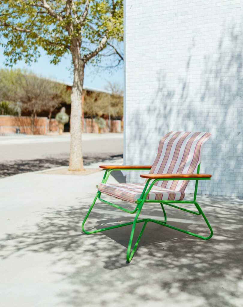 A striped outdoor chair with green metal legs and wooden armrests sits on a sunlit sidewalk near a white brick wall and a tree.