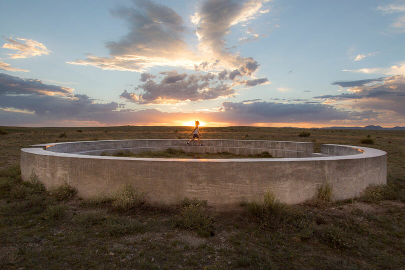 Joey Benton walks along a large circular concrete art installation in an open landscape at sunset, with clouds and distant mountains in the background.