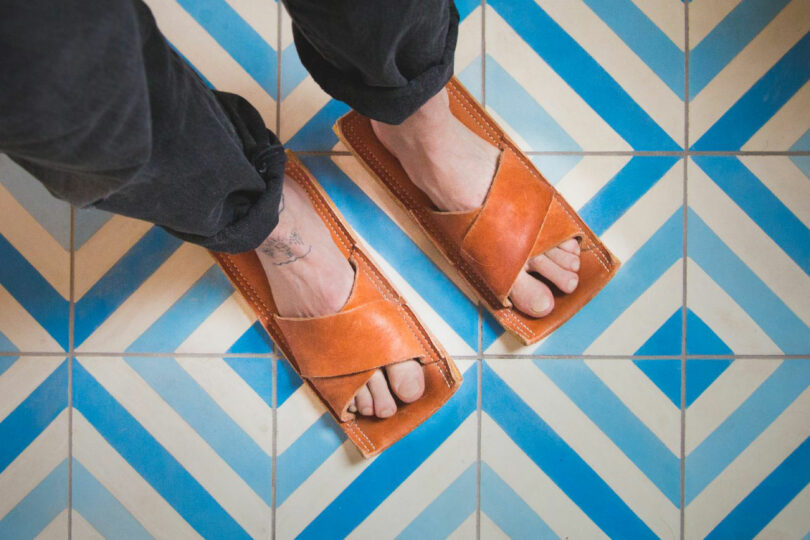 Person wearing brown leather sandals with square soles standing on blue and white geometric patterned tiles.