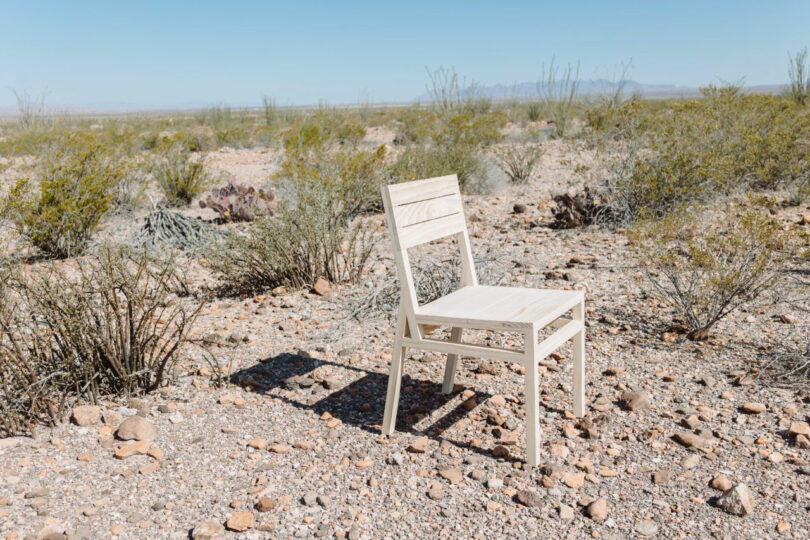 A simple wooden chair is placed on rocky, dry ground in a desert landscape with sparse vegetation under a clear blue sky.