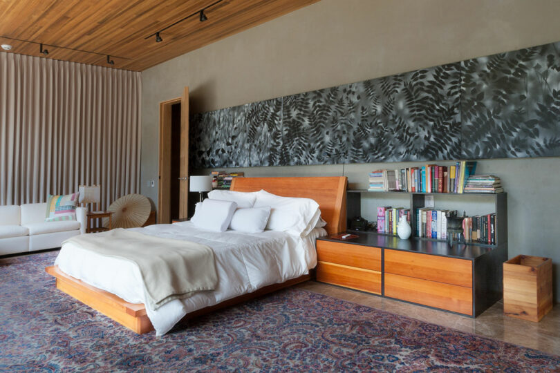 Modern bedroom by Joey Benton featuring a wooden bed, white bedding, two-tone nightstands, bookshelves, patterned rug, and abstract art above the headboard. A white sofa and accent chair complete the cozy corner.