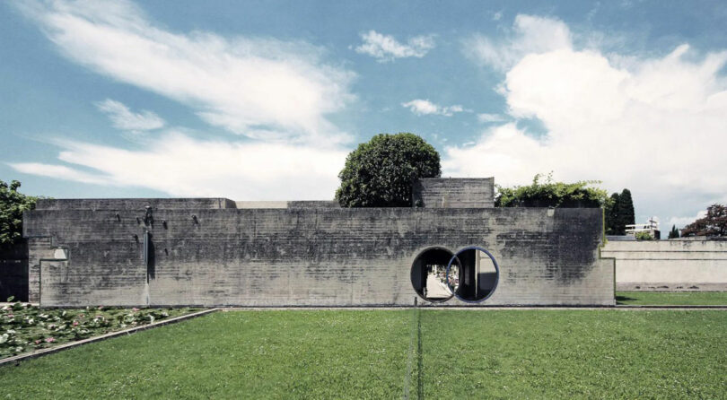 A modern concrete building by Tyler Kobick, featuring circular openings, stands on a green lawn under a blue sky with scattered clouds.
