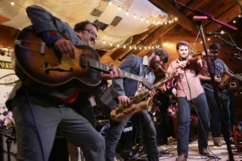 Tyler Kobick leads a band on stage; one plays guitar, another saxophone, a third plays fiddle, and a fourth plays accordion under string lights in a rustic setting.