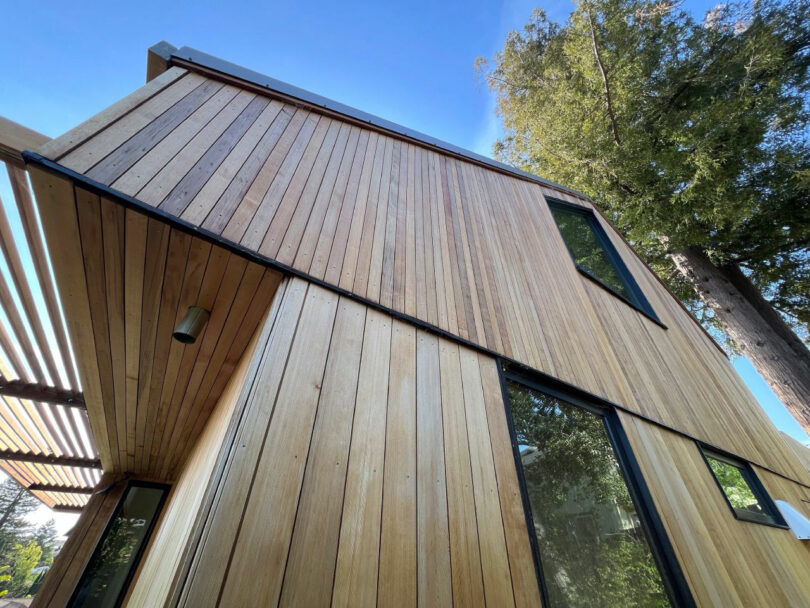 A modern house exterior by Tyler Kobick features vertical wooden siding, large windows, and a slatted pergola, set against a clear blue sky and tall trees.