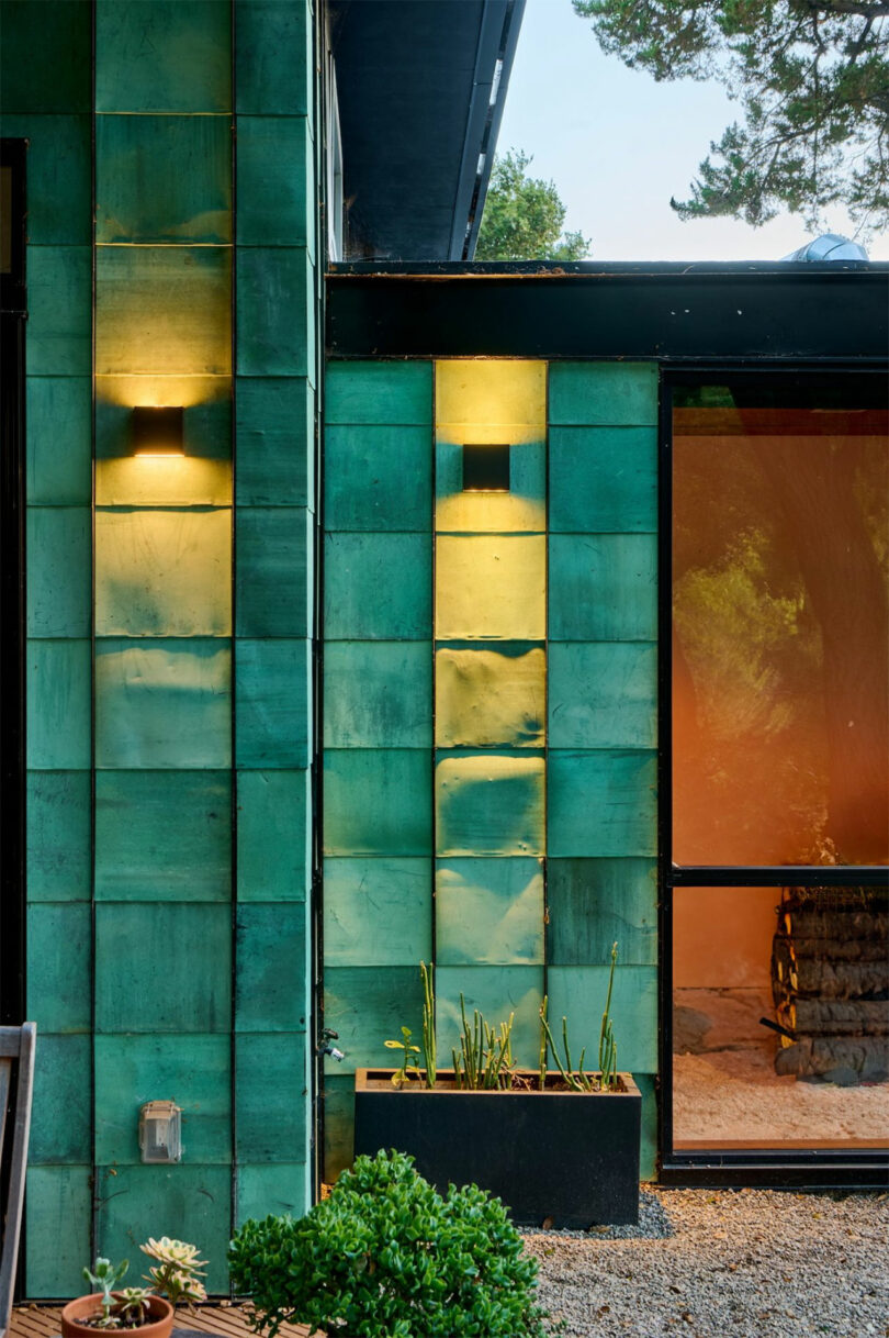 Exterior wall with large green ceramic tiles by Tyler Kobick, modern wall lights, a black planter with plants, gravel ground, and a glass door leading inside.