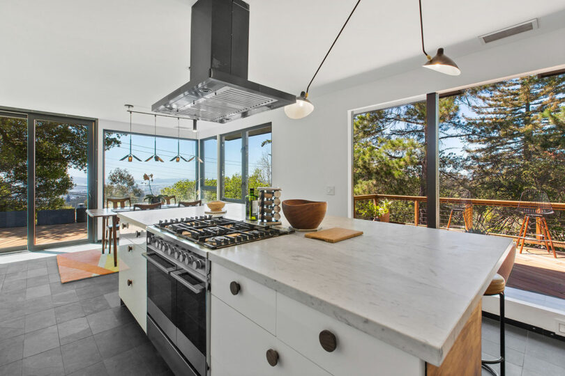 Modern kitchen designed by Tyler Kobick with a marble island, gas stove, and large windows overlooking a deck and trees. Ample natural light fills the space.