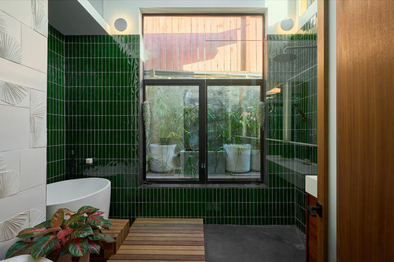 Modern bathroom with green tile walls, a freestanding bathtub, wooden bench, potted plant, and natural light streaming in—a serene space inspired by Tyler Kobick’s signature style.