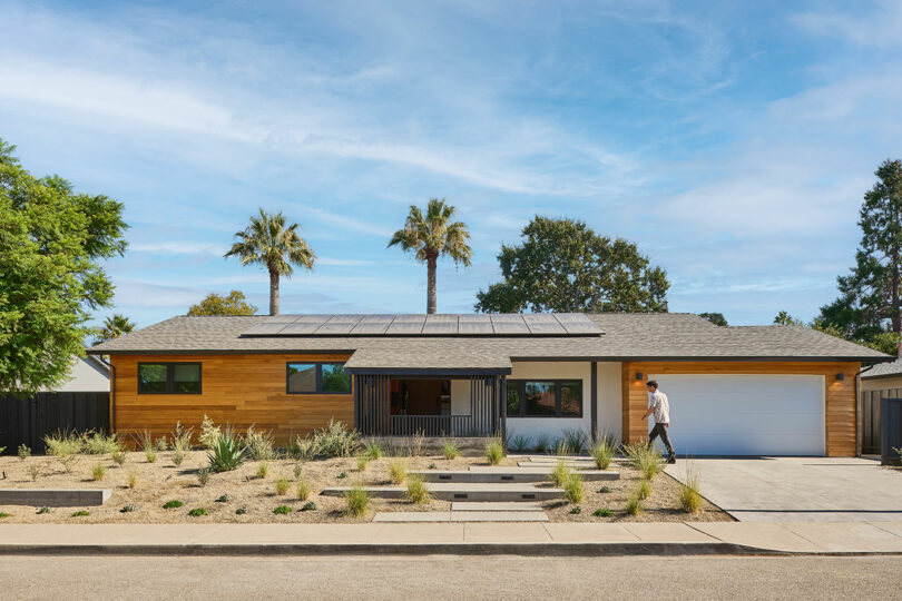 A single-story modern house with solar panels on the roof, a xeriscape front yard, and a person walking on the pathway toward the garage showcases Tyler Kobick's signature sustainable design.