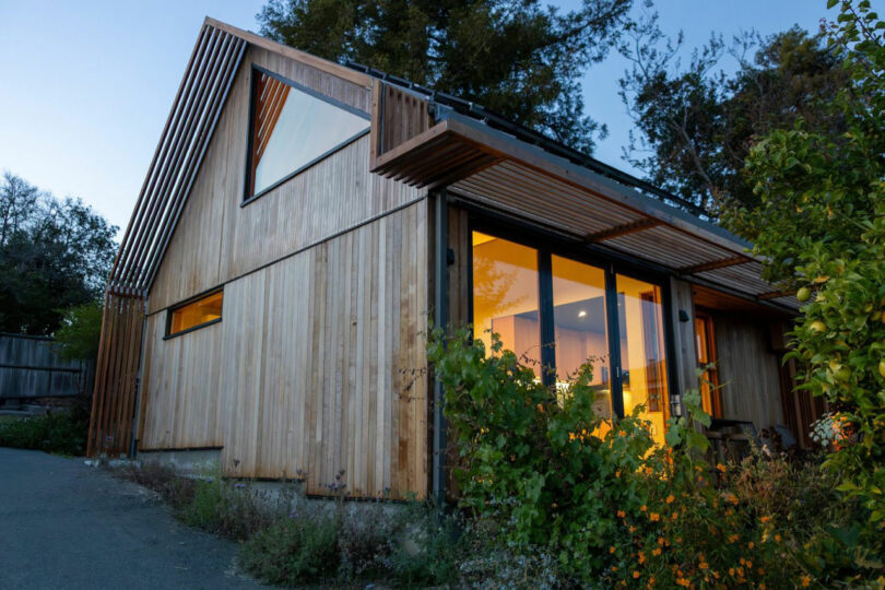 Modern wooden house with large windows and warm interior lighting, designed by Tyler Kobick, surrounded by greenery in the evening.