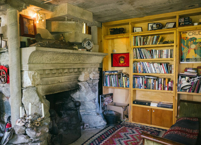 A stone fireplace sits beside a wooden bookcase filled with books and decor items in a cozy, rustic room designed by Tyler Kobick, featuring a patterned rug and eclectic furnishings.