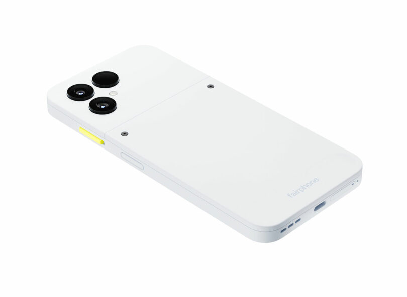 A white Fairphone smartphone is shown facing down, highlighting its triple rear cameras, yellow side button, and Fairphone branding near the bottom—showcasing Fairphone’s signature sustainable design.