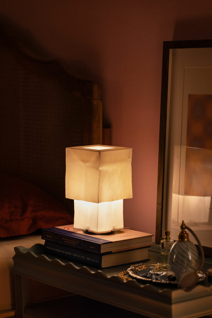 Eine quadratische Tischlampe mit einem weichen, weißen Schatten sitzt auf einem Haufen Bücher neben einem gerahmten Bild, Parfümflaschen und einem dekorativen Tablett am Nachttisch.
