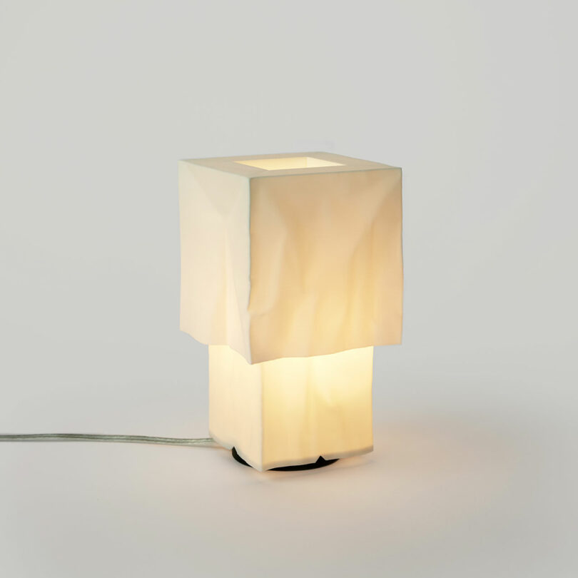 Moderne Tischlampe mit weißem, zerknittertem quadratischem Farbton, aus dem einfachen Hintergrund weich warm und sichtbares Netzkabel.