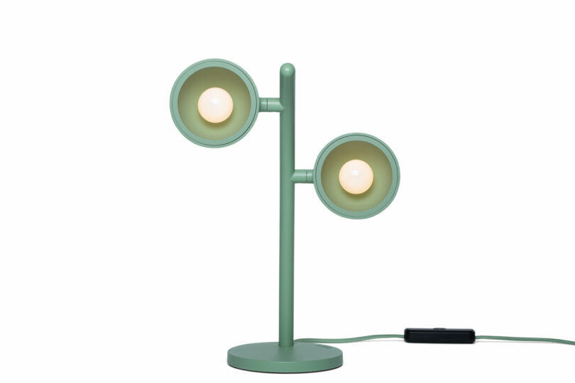 Green desk lamp with two round light fixtures on a single vertical stand, each facing a different direction, and an attached power cord with a switch.