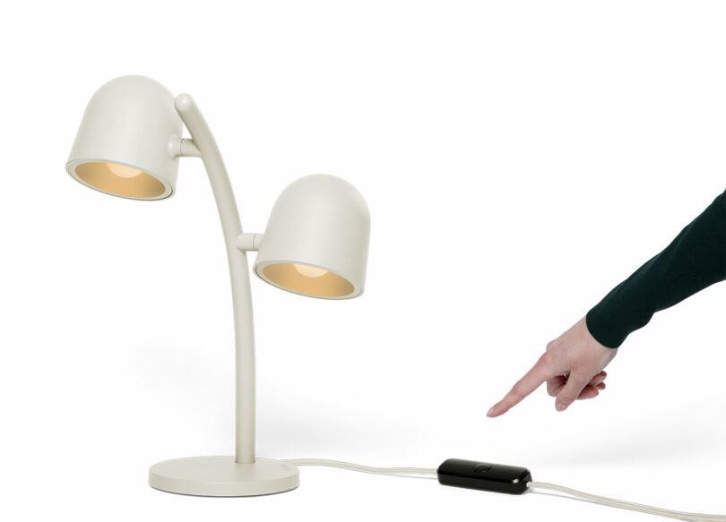 A person points to a switch on the power cord of a white dual-headed desk lamp against a white background.