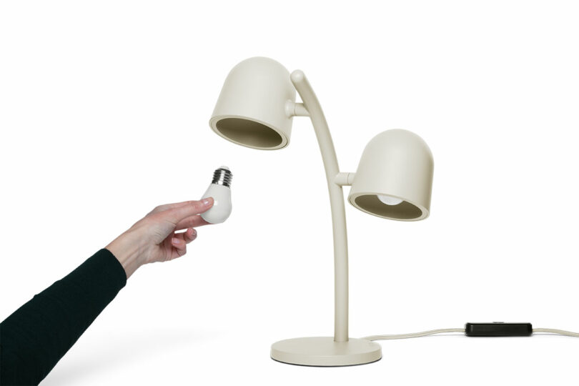 A hand is holding a light bulb next to a modern, double-headed beige desk lamp on a white background.