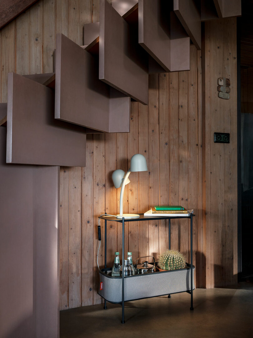 A small console table with a lamp, books, bottles, a cactus, and a bowl sits against a wooden wall under a modern staircase.