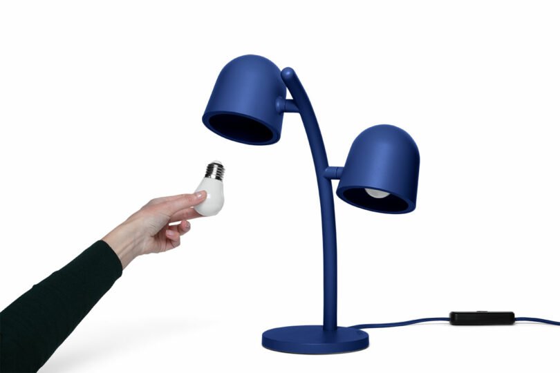 A hand holds a light bulb near a blue double-headed desk lamp against a white background.