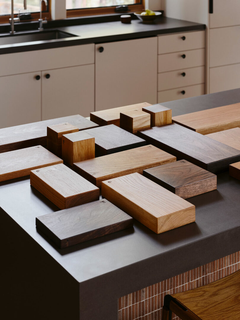 Auf einer dunklen Kücheninsel mit einem leichten Schrank und einer Spüle im Hintergrund sind verschiedene rechteckige Holzproben angeordnet.