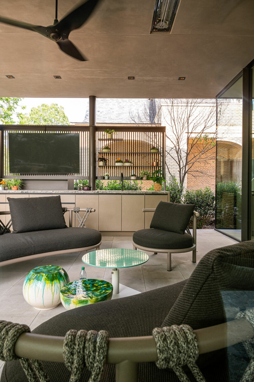 Modern outdoor patio with dark cushioned seating, round glass coffee table, decorative poufs, wall-mounted TV, and a vertical garden on the fence in the background.