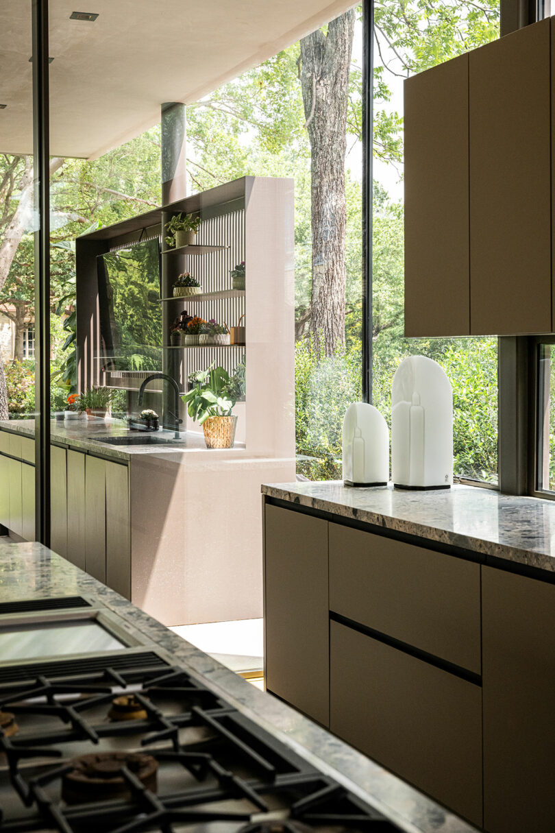 Modern kitchen with large windows, beige cabinetry, marble countertops, and a view of greenery outside. Decorative white vases and plants are placed on the counters and shelves.